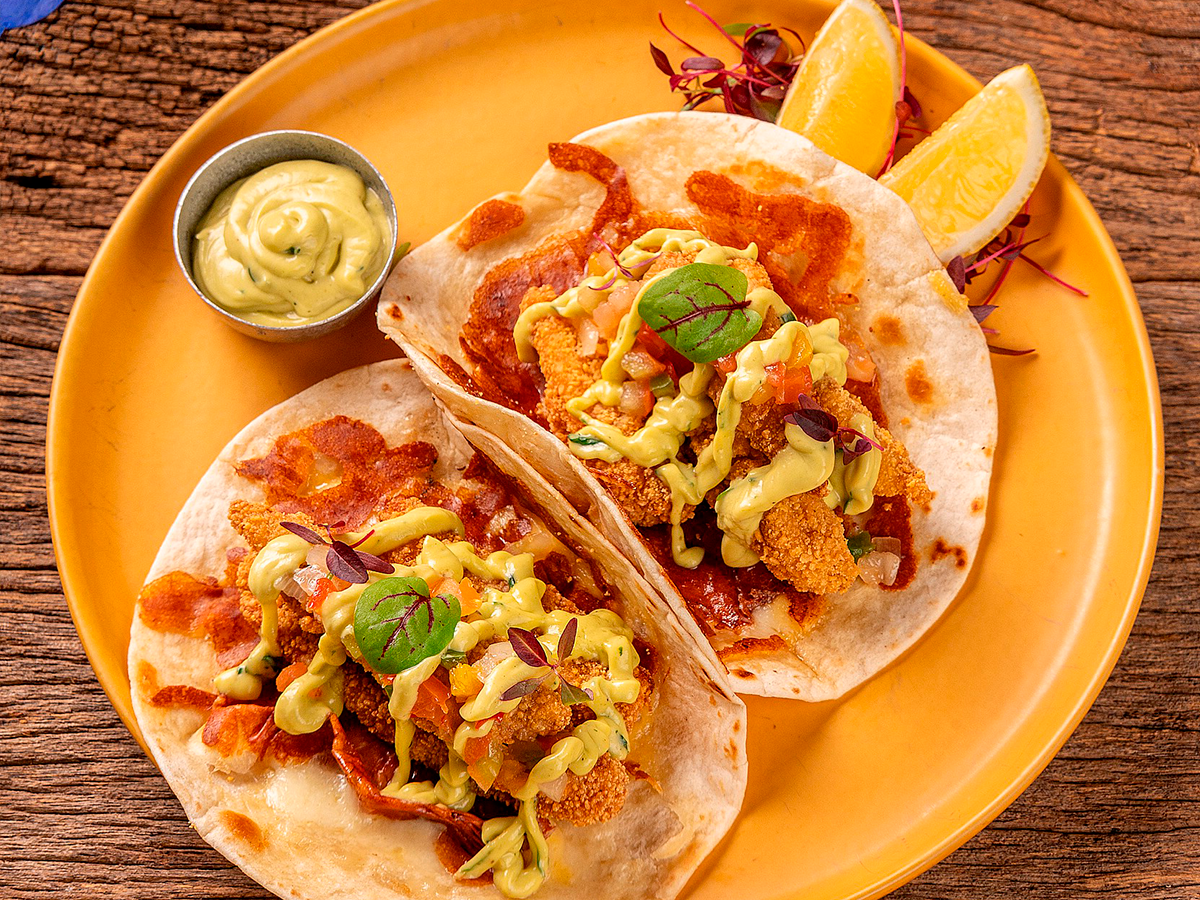 Tacos servidos em um prato laranja com molho para mergulhar e fatias de laranja, decorados com coentro.