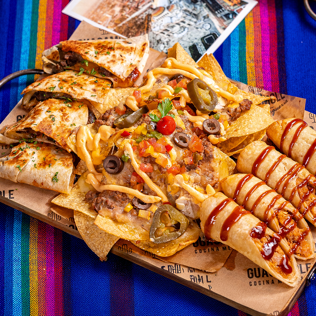 Uma bandeja com uma variedade de comidas de rua mexicanas: tacos, nachos e tortillas enroladas com molhos sobre uma toalha de mesa listrada e colorida.