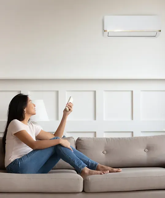 Woman on couch using remote to control wall-mounted air conditioner in a white-walled room.