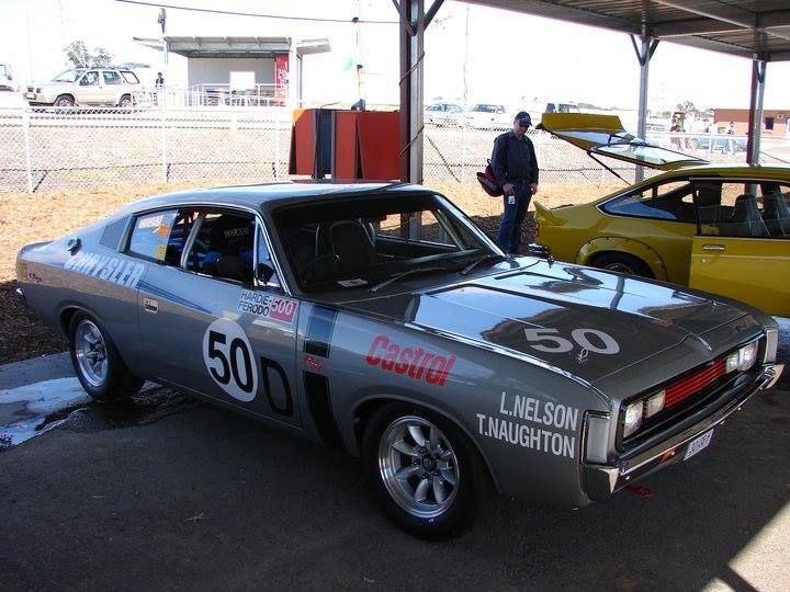 Gray race car number 50 with sponsor decals parked under a canopy, yellow car and person in background