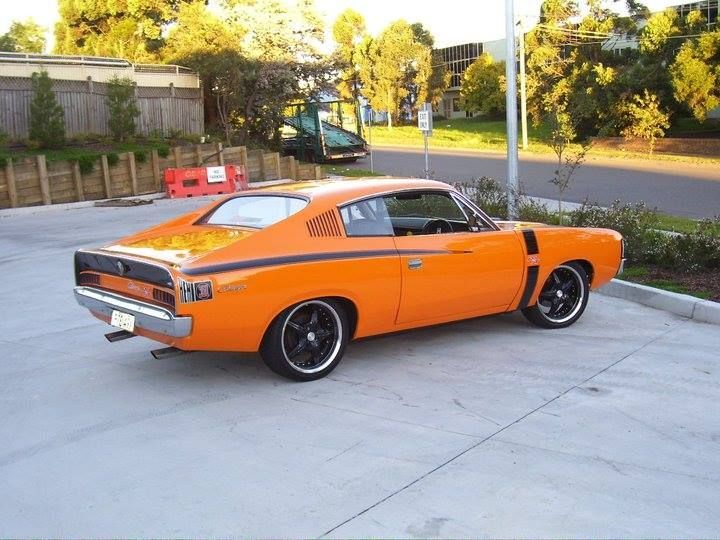 Orange classic muscle car parked on a concrete driveway near trees and a road