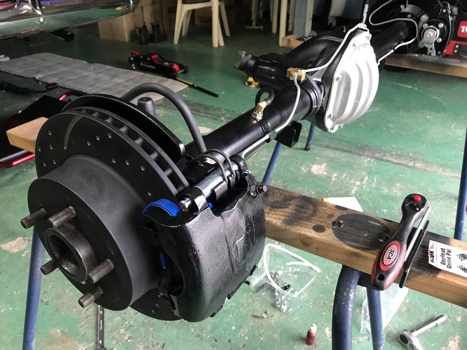 Rear disc brake assembly mounted on a black axle housing in a workshop, with tools and a workbench nearby