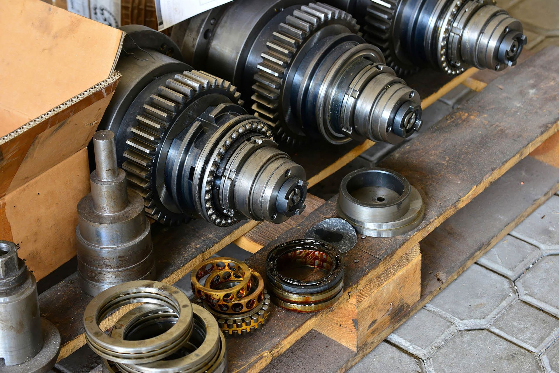 Mechanical gears and metal parts arranged on a workshop floor beside a wooden crate.