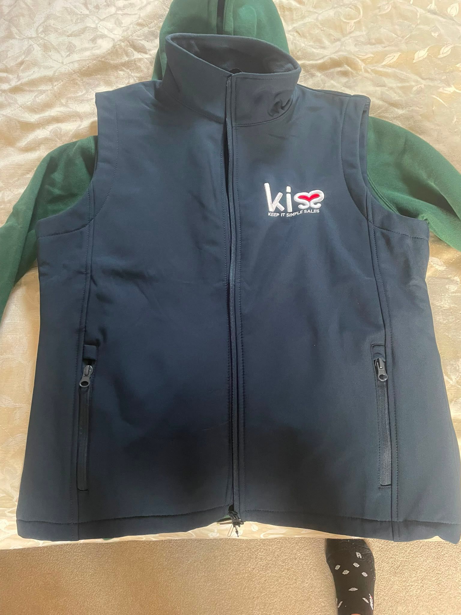Black sleeveless vest with green sleeves underneath, laid flat on a beige fabric surface.