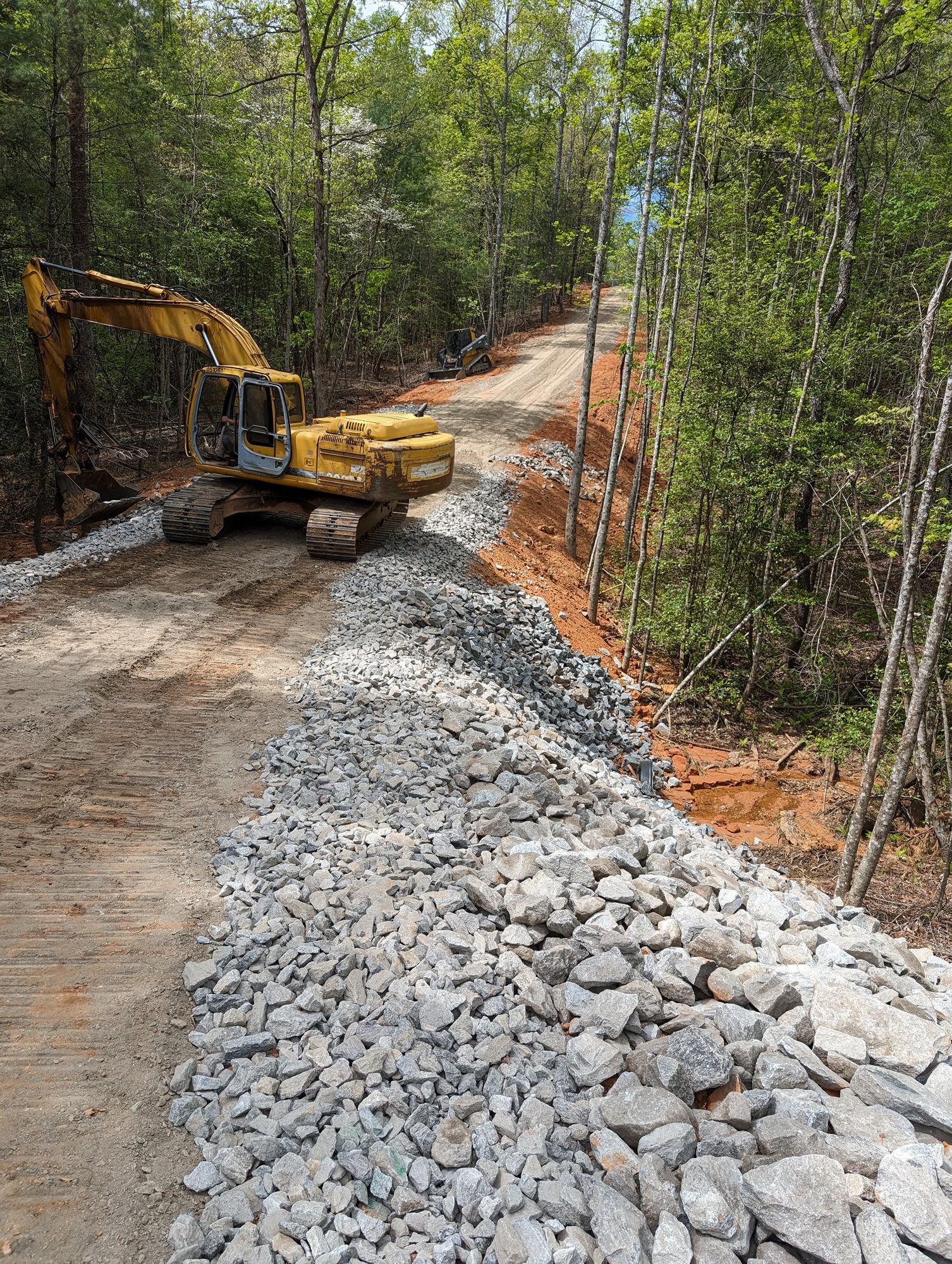 Yellow excavator on a forest road beside a pile of gray rocks and orange erosion fence