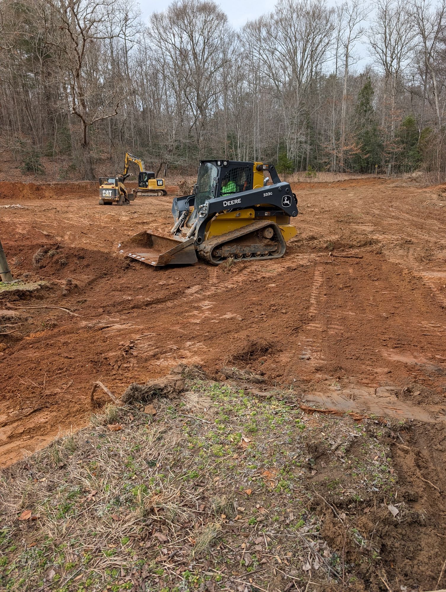 Tracked skid steer grading red dirt in a cleared wooded area, with another machine in the background.