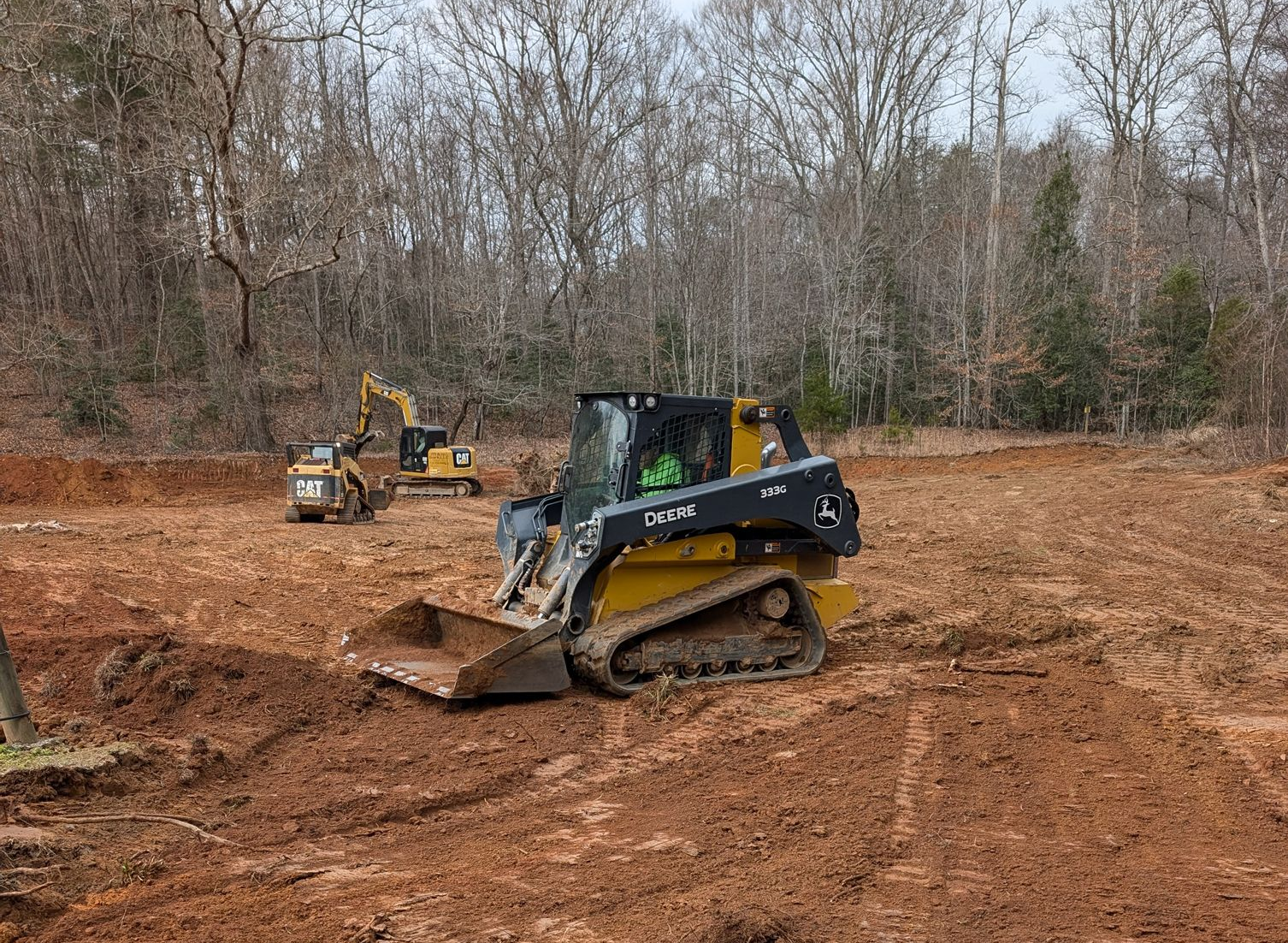 A black-and-yellow skid steer on muddy ground with a small excavator in the background among bare trees.