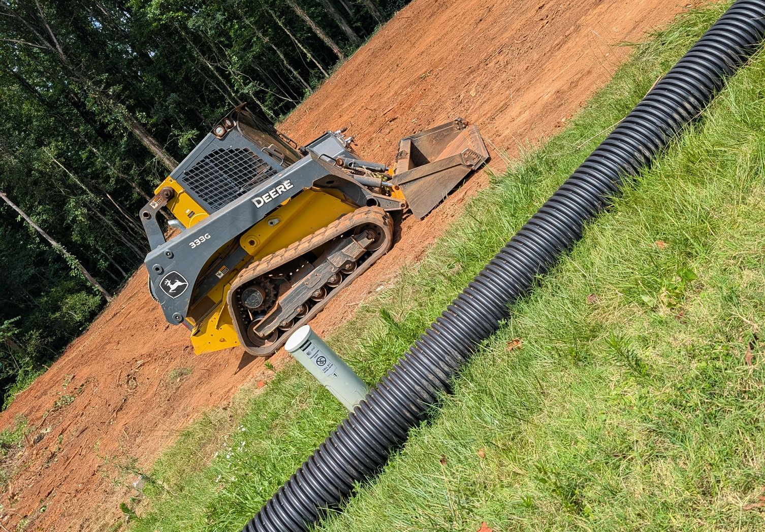 Yellow skid steer on dirt beside black drainage pipe and grassy slope, outdoors at a construction site