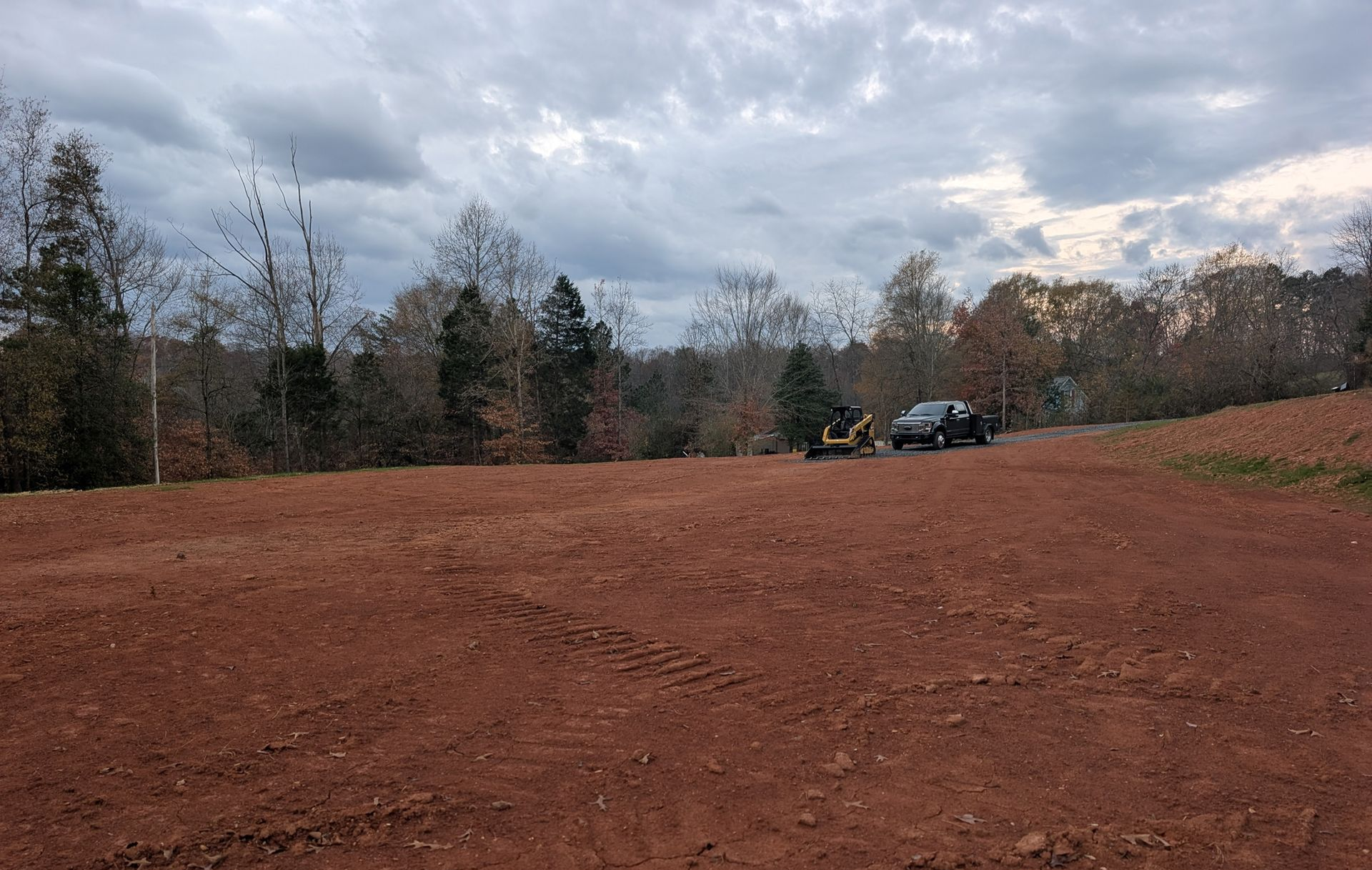 Dirt clearing beside a wooded edge under cloudy sky, with a parked truck near the trees