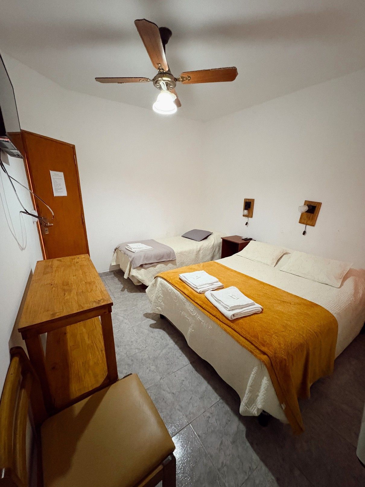 Habitación pequeña con cama, mesita de noche, silla, escritorio y ventilador de techo; paredes blancas y detalles de madera.