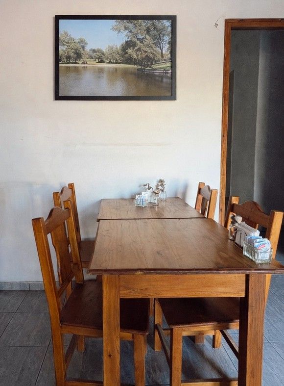 Mesa de comedor con sillas de madera en una habitación sencilla, con un cuadro de paisaje en la pared.