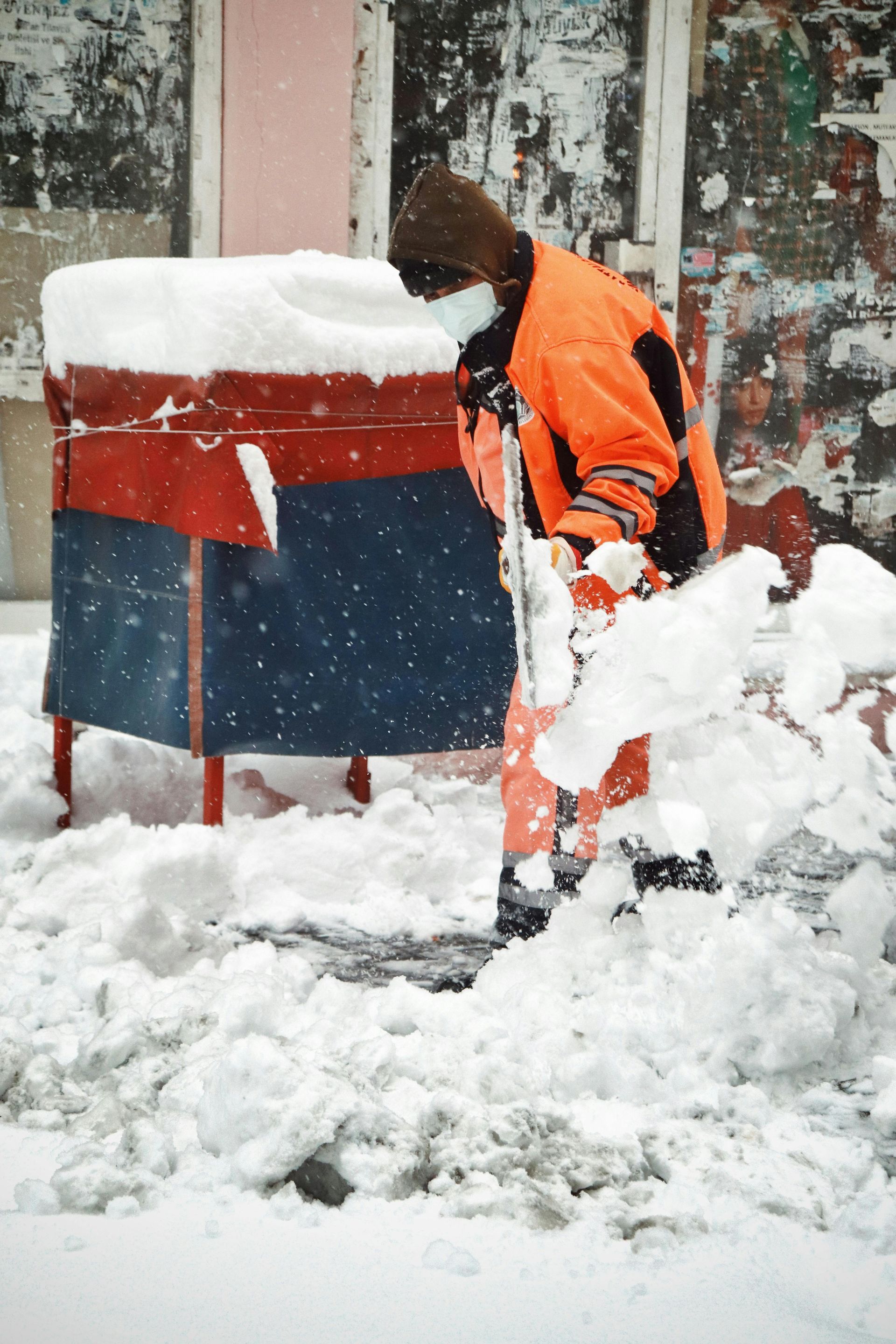 A man wearing a mask is shoveling snow in front of a trash can
