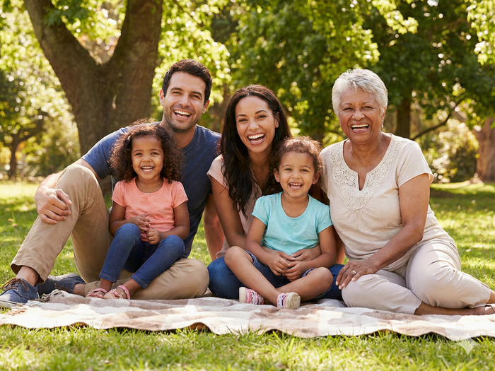 A multigenerational family smiles together while sitting on a blanket on the grass in a park.