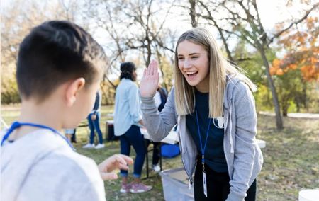 A volunteer high-fives a child outdoors at a park, both smiling and interacting during a community event.