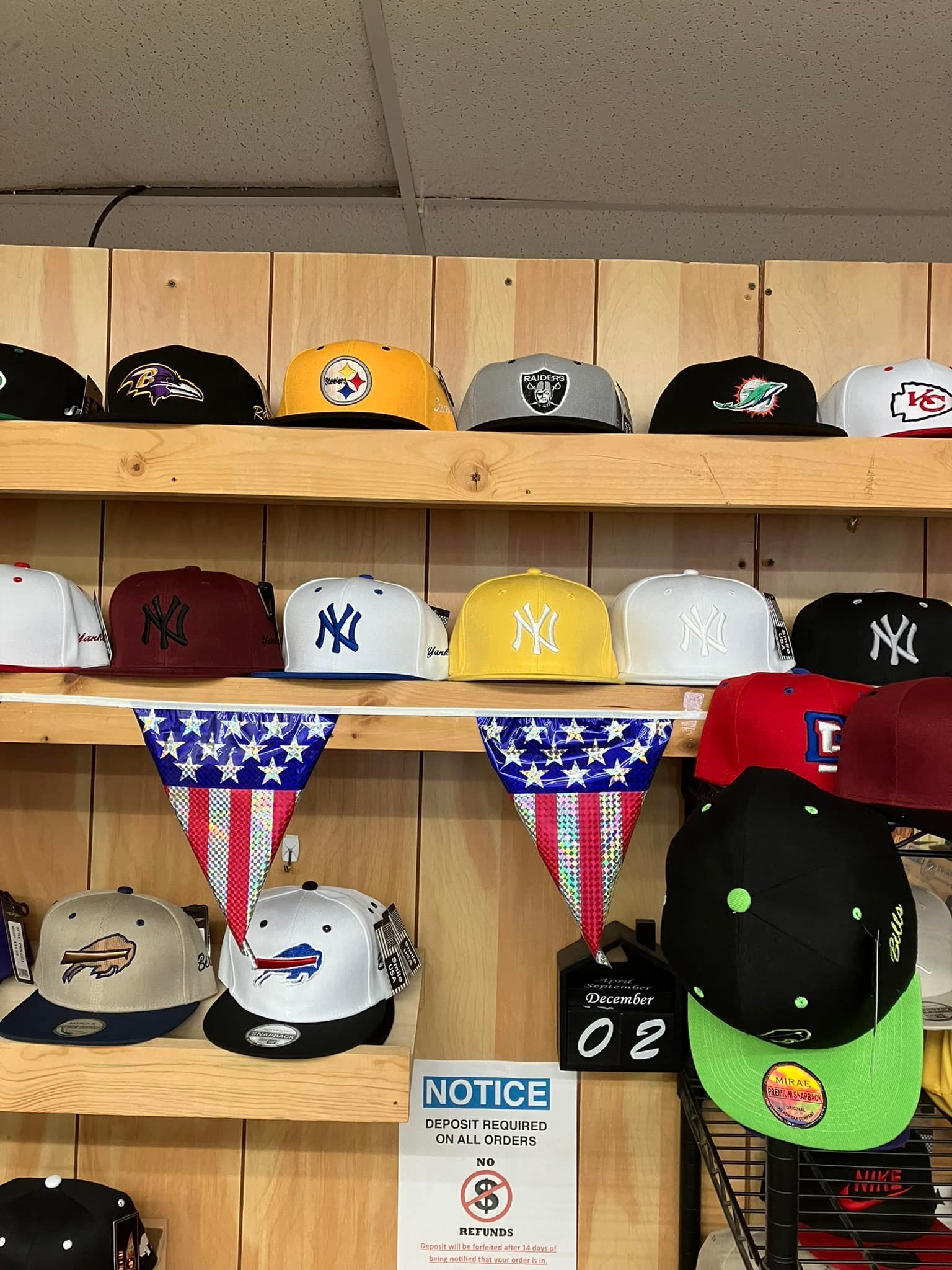 Multiple shelves display various sports team baseball caps in a retail store, with American flag bunting hung below.