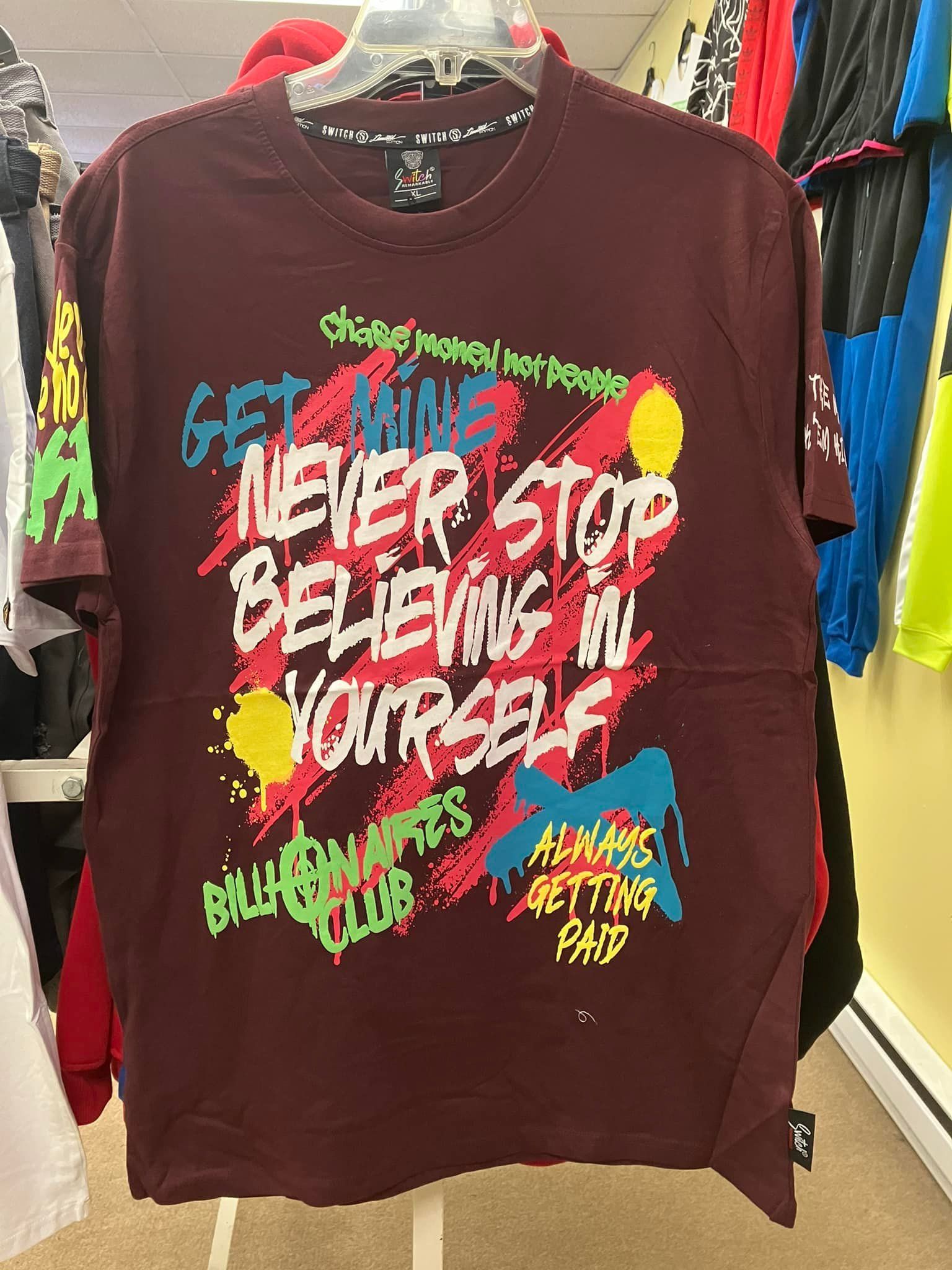 A maroon graphic t-shirt hanging on a rack features colorful, spray-paint style text reading 