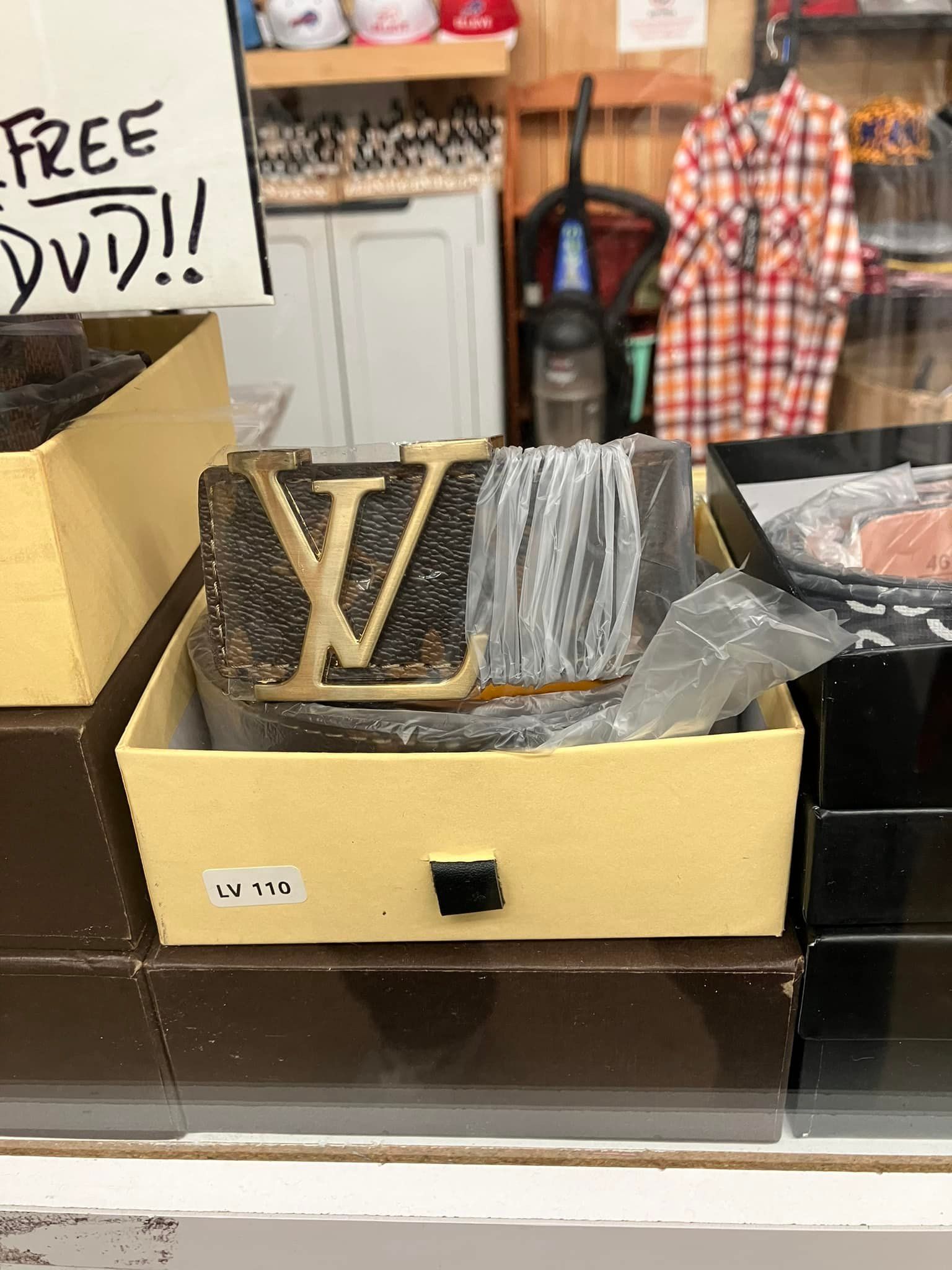 A Louis Vuitton belt with a gold-tone logo buckle, wrapped in protective plastic inside a tan gift box on a shelf.