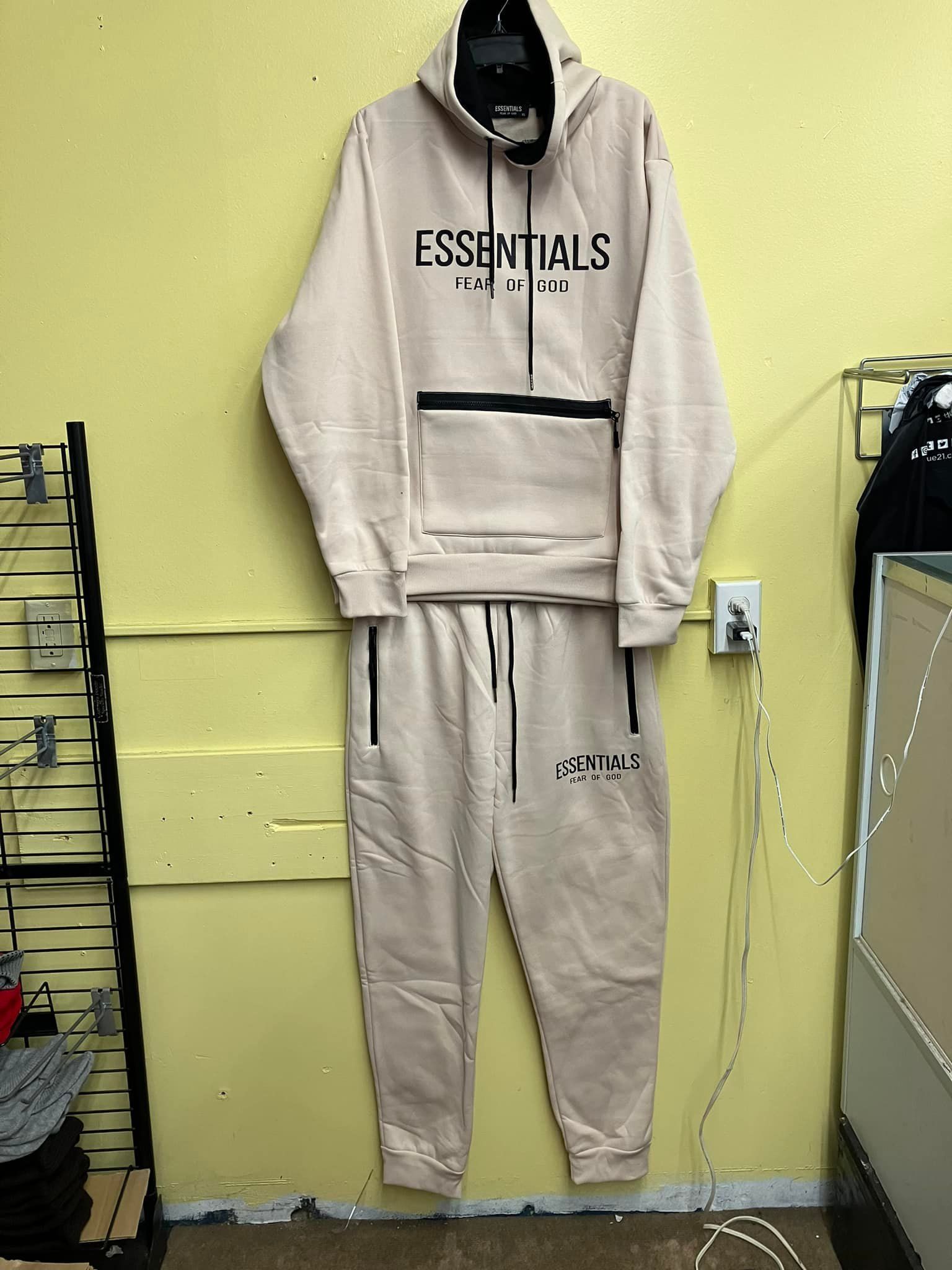 A beige Essentials hoodie and matching jogger set hanging against a pale yellow wall.