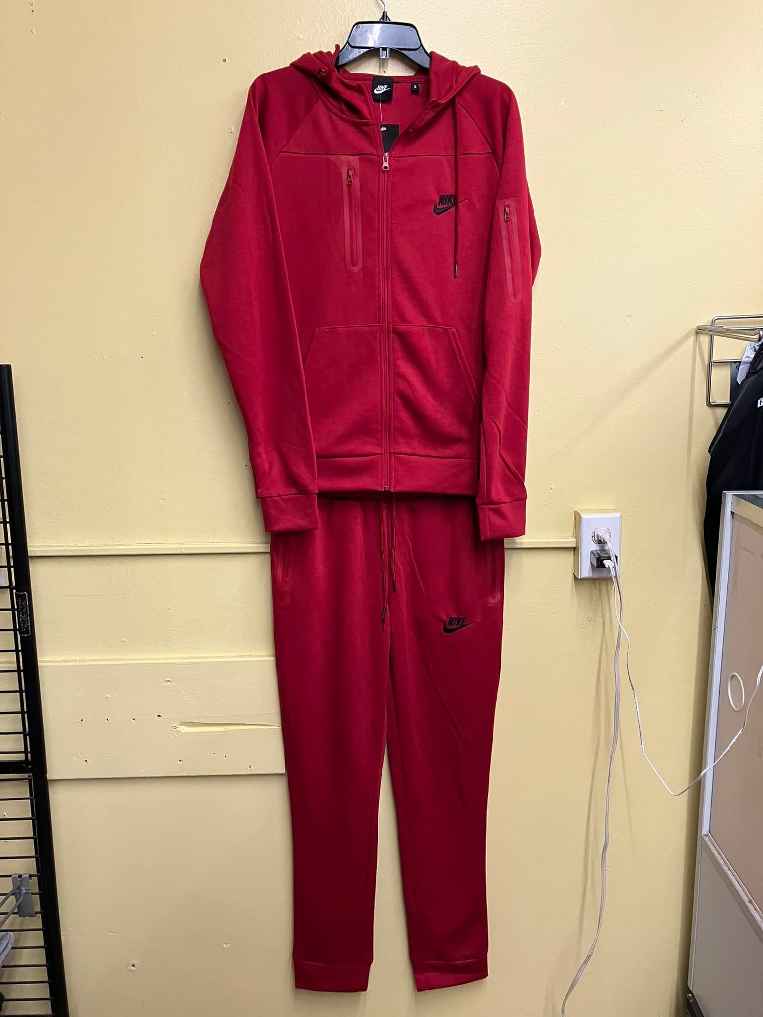 A bright red matching tracksuit consisting of a zip-up hoodie and sweatpants, hanging against a plain yellow wall.