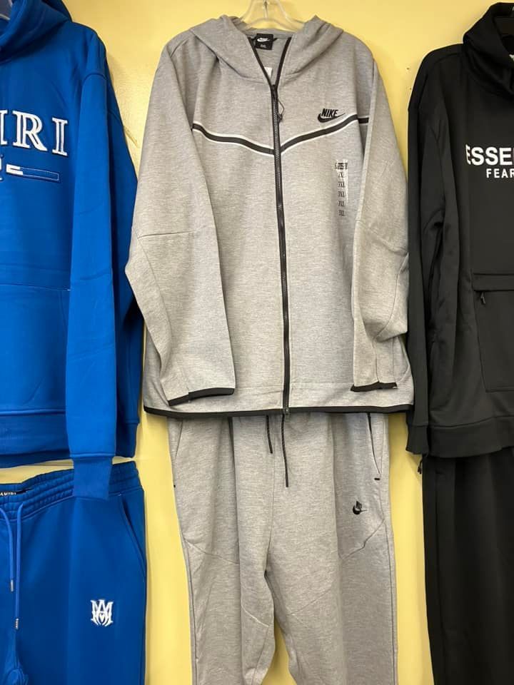 A gray Nike Tech Fleece hooded tracksuit displayed on a hanger between a blue and a black hoodie against a yellow wall.