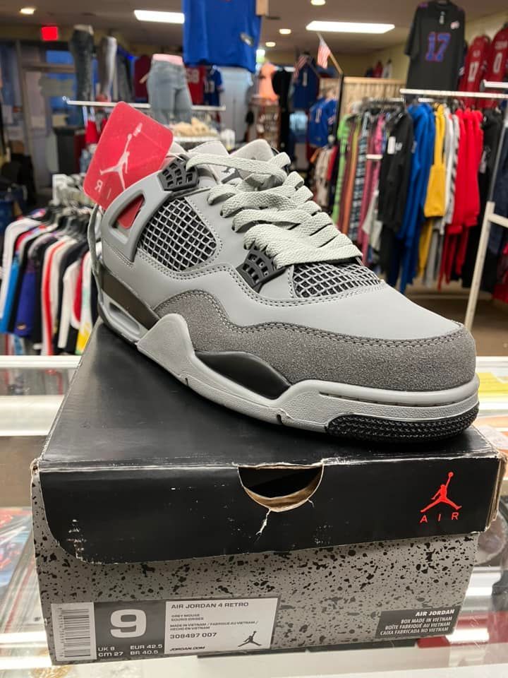 A grey and black Jordan 4 sneaker sits on its cardboard box inside a retail store with clothing racks in the background.
