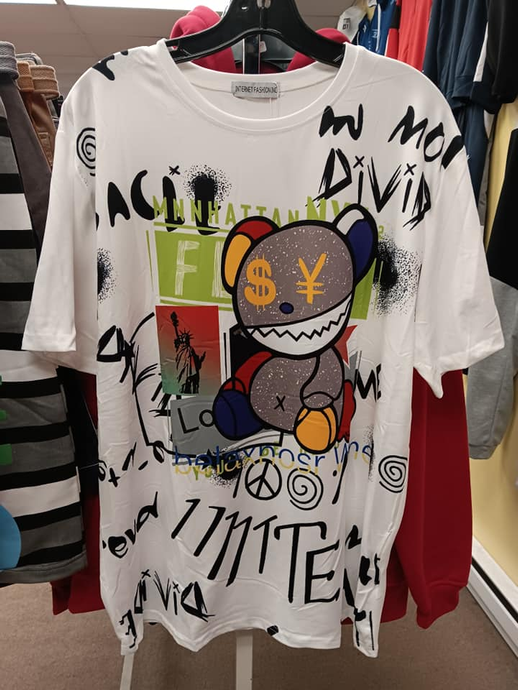 A white T-shirt on a hanger with a graphic of a teddy bear with money symbols and black graffiti-style text patterns.