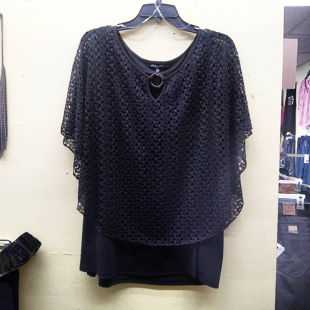 A black, short-sleeved top with a layered, lace-patterned poncho overlay and a decorative cut-out at the neckline.