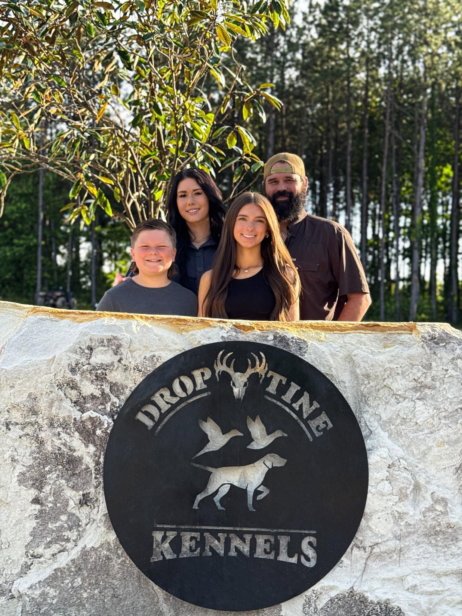 Four people posing behind a stone sign for “Drop Line Kennels” outdoors in the sun