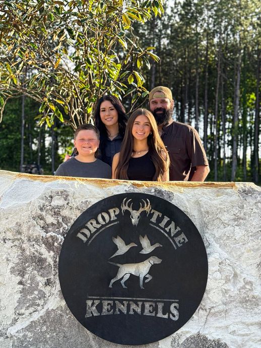 Four people posing behind a stone sign for “Drop Line Kennels” outdoors in the sun