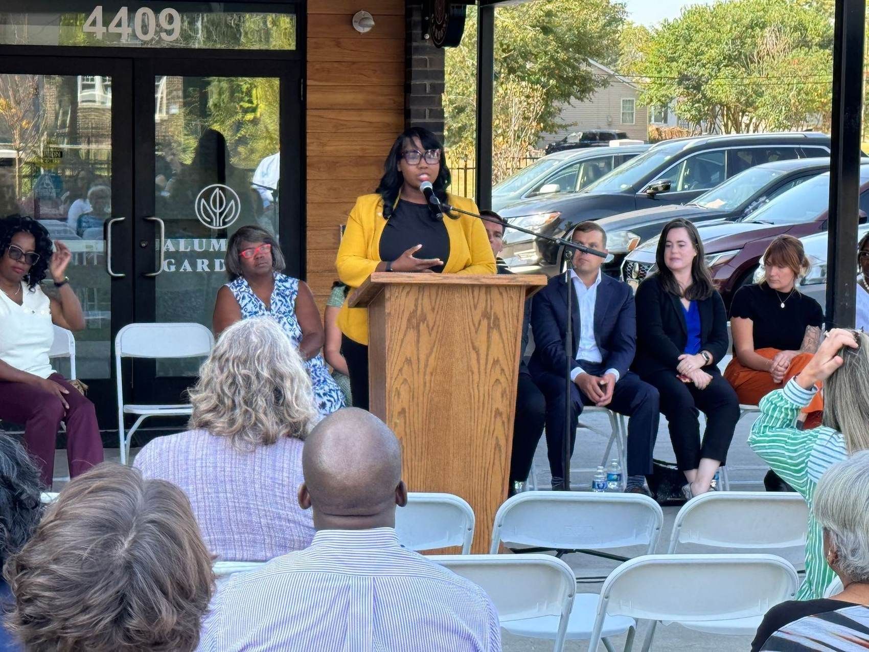 A speaker addresses seated guests at the Alumnus Gardens opening ceremony, outside the finished building at number 4409.