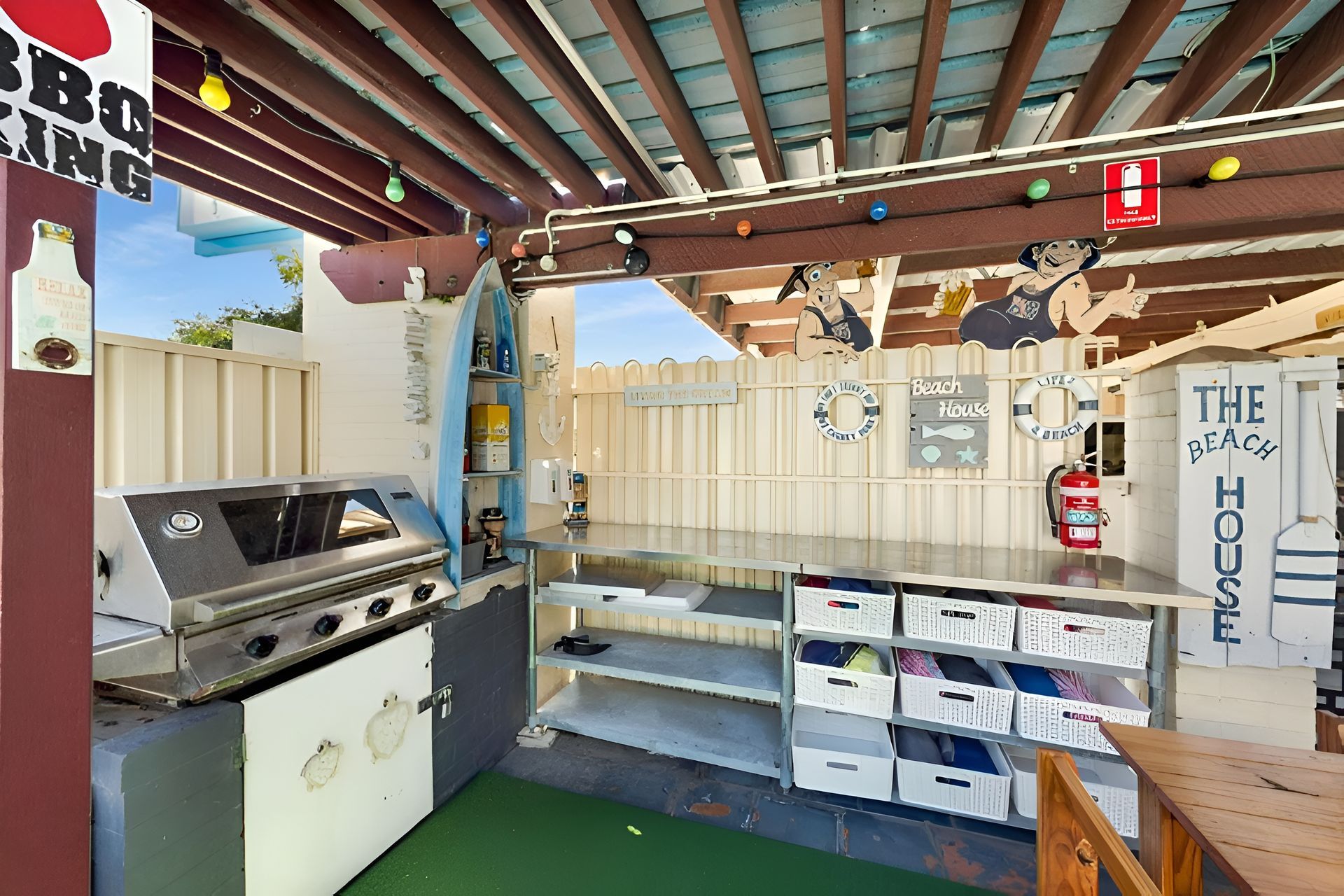 Covered outdoor patio with a grill, shelves, storage bins, string lights, and a “The Home” sign.