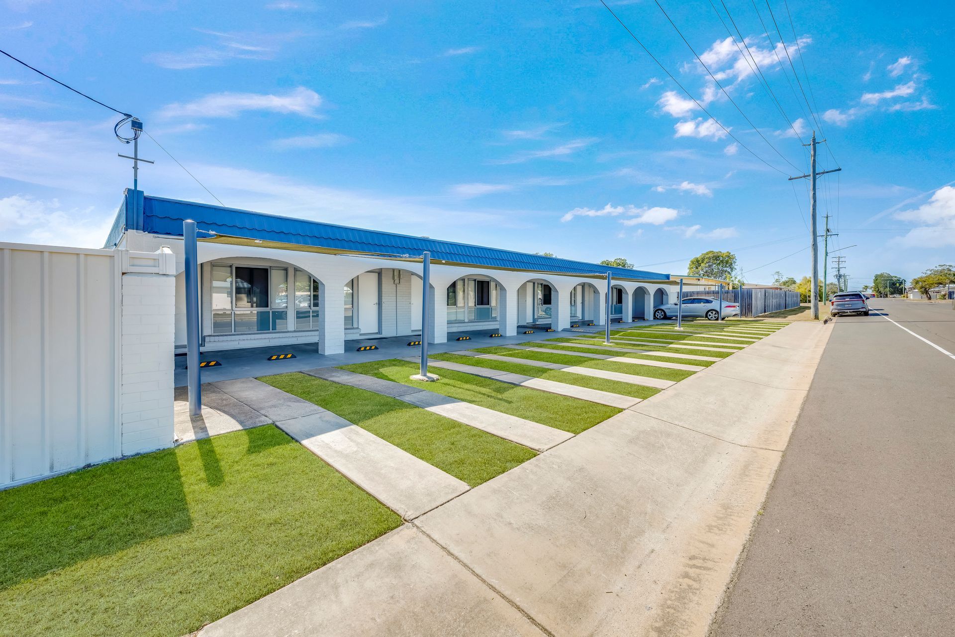 Covered walkway beside a grassy courtyard with white arches, striped paving stones, and a parked car