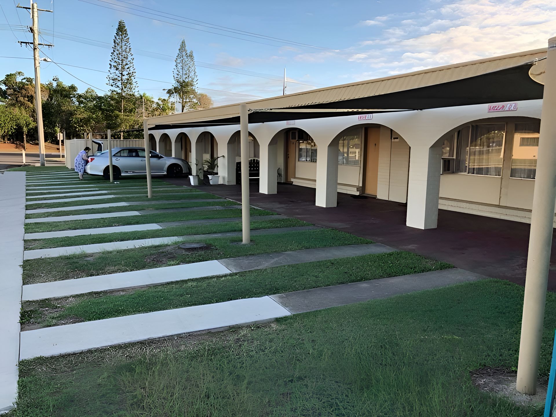 Covered walkway beside a grassy courtyard with white arches, striped paving stones, and a parked car