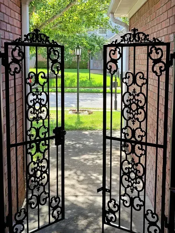 Open black wrought-iron gate leading to a sunny front yard with trees and a sidewalk.