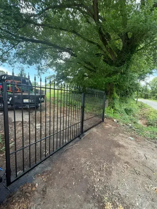 Black metal gate beside a dirt path under a large green tree, with a truck parked behind it.