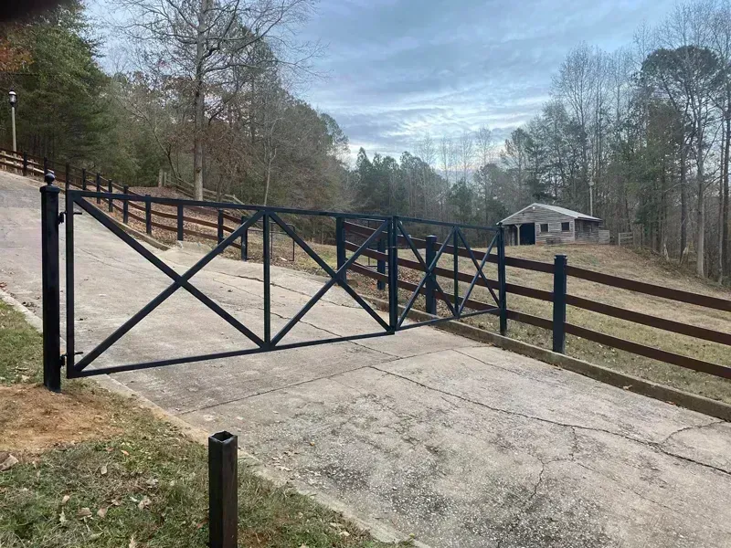Black metal gate across a sloped concrete driveway in a wooded rural area