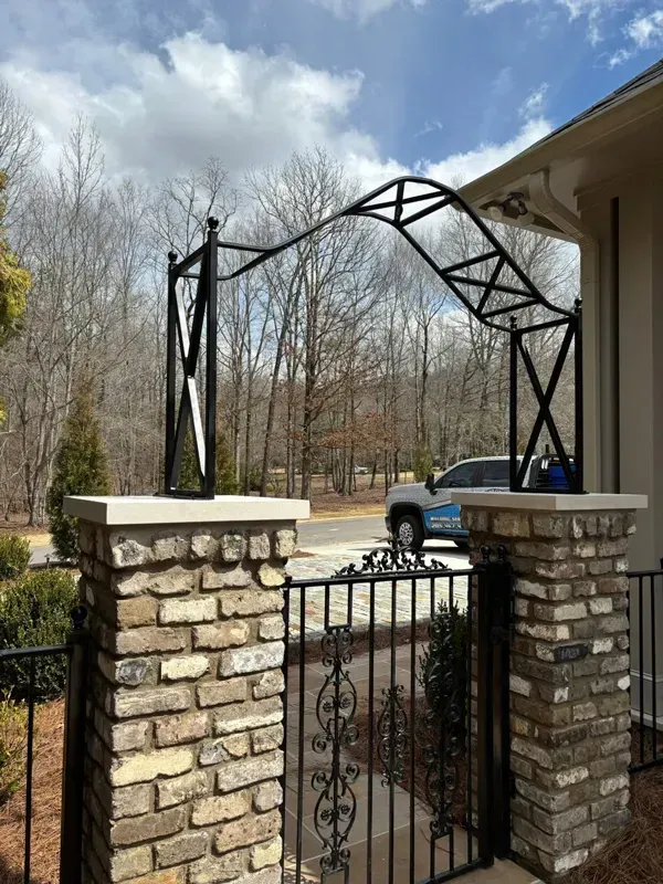 Stone gate pillars and black metal archway over a driveway, with a blue pickup truck and leafless trees beyond.