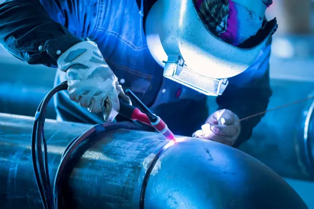 Welder in blue protective gear working on a large metal pipe with sparks and arc light