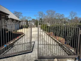 Open metal gate at the entrance to a driveway beside a house and landscaped shrubs.
