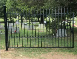 Black iron gate in front of a cemetery with headstones and trees in the background.
