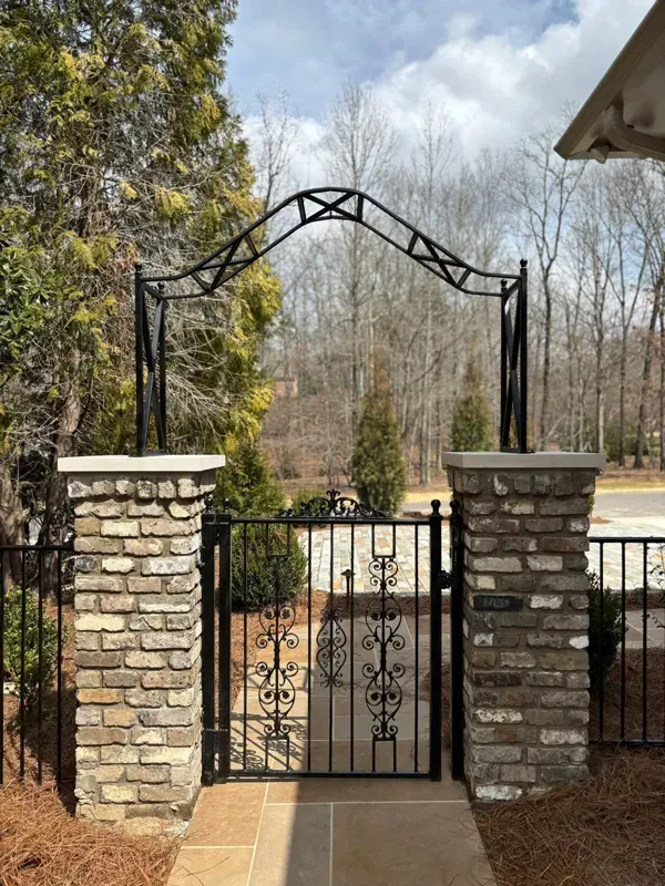 Wrought-iron gate between stone pillars under an arched trellis, opening to a wooded path