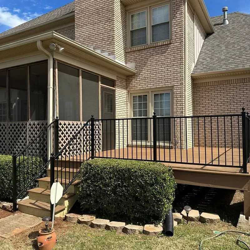 Brick house back patio with black metal railing, screened porch, and trimmed shrubs under a sunny sky