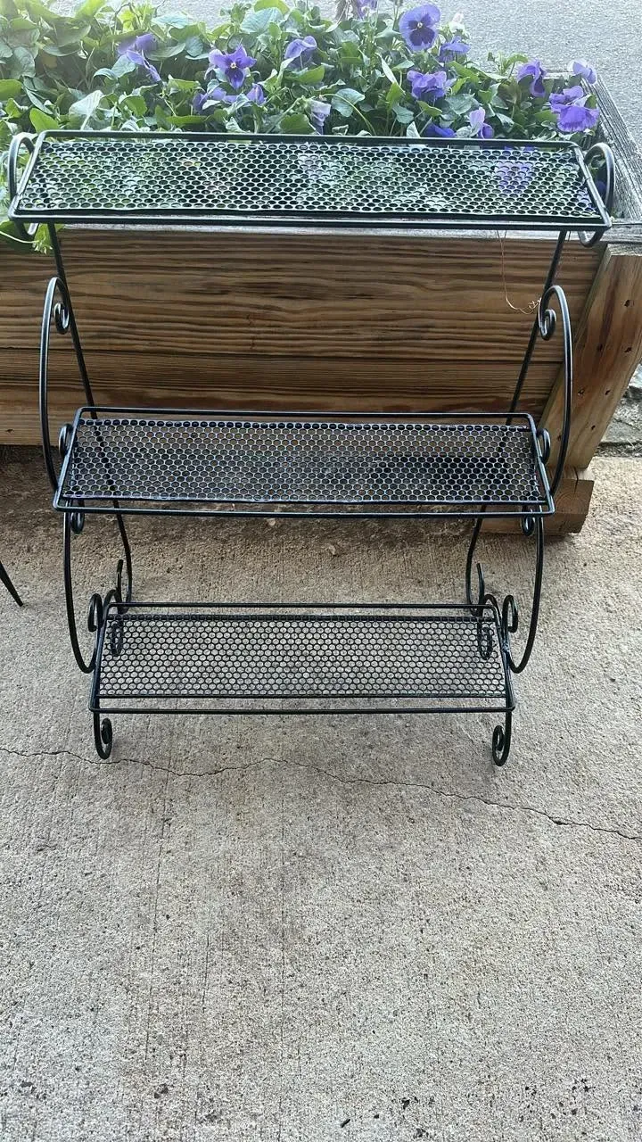 Three-tier black wire plant stand with purple flowers on top, in front of a wooden planter box on a patio.
