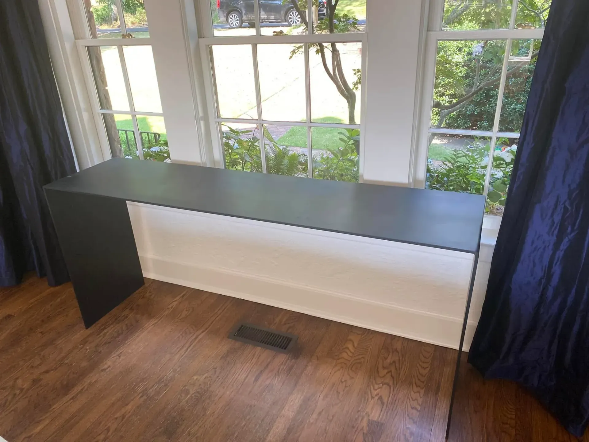 Black desk against a window with white blinds and blue curtains on a hardwood floor