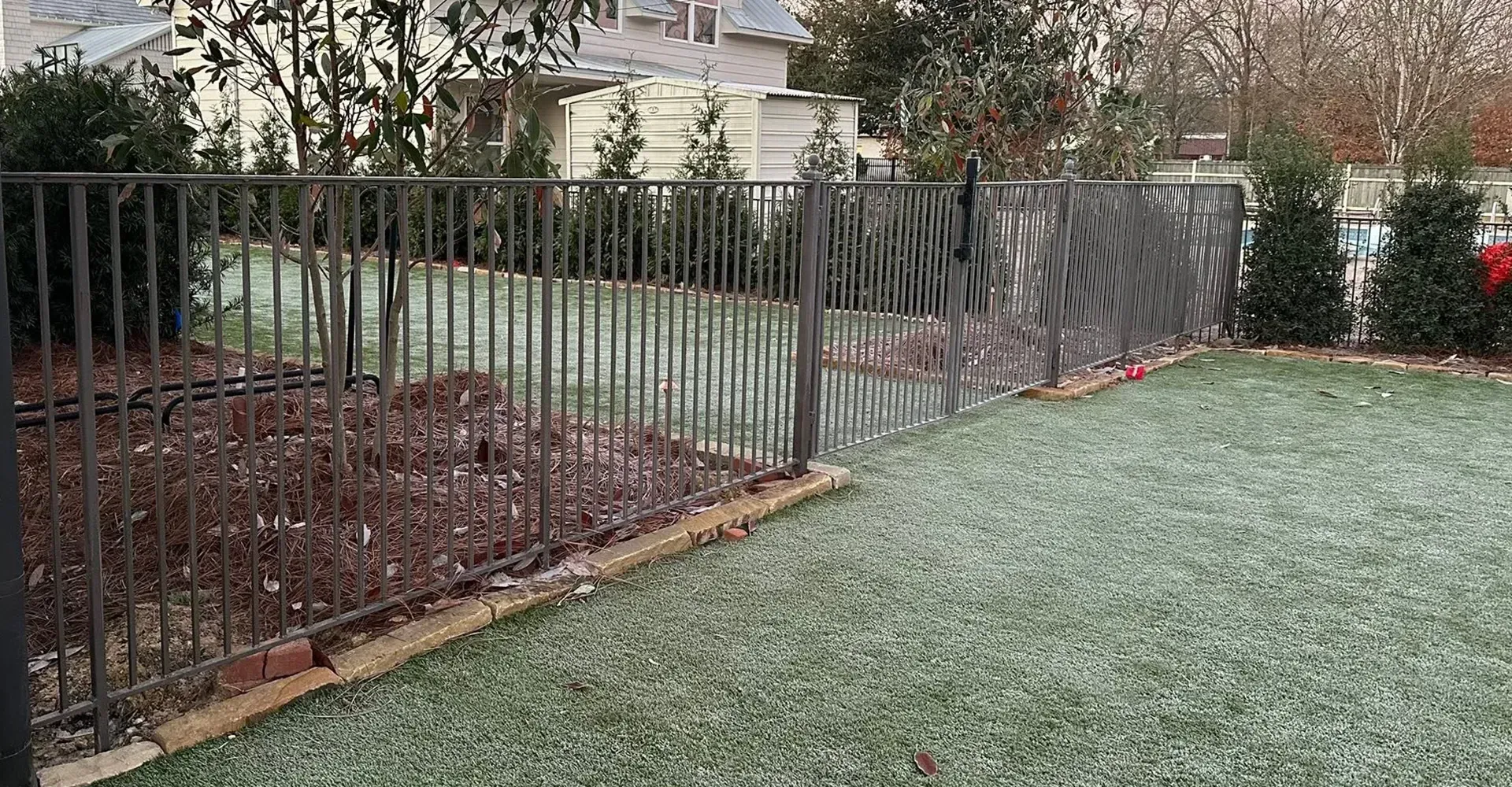 Black metal fence beside a frosty backyard lawn with shrubs and a house in the background
