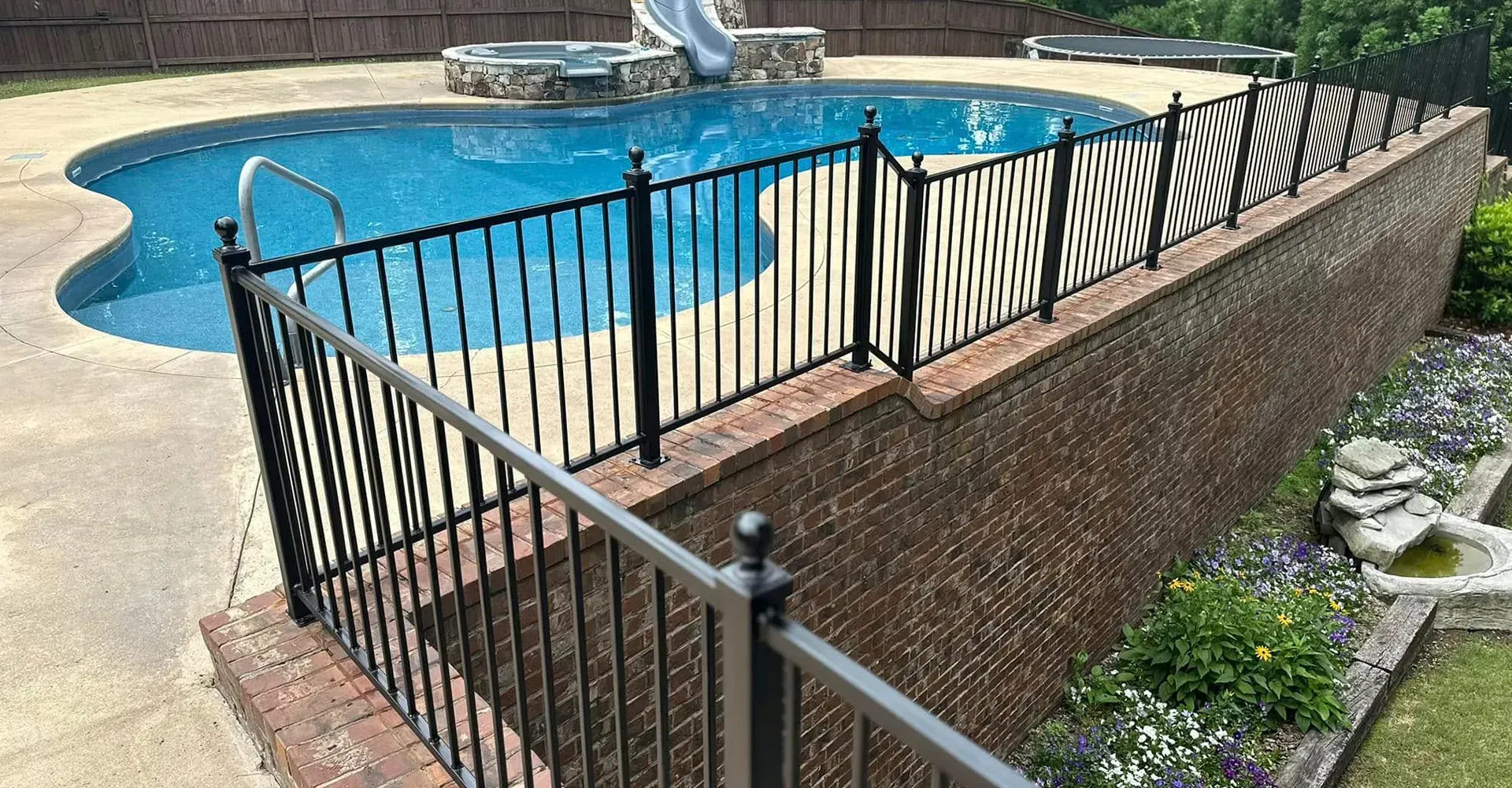 Black metal fence around a raised pool deck beside a blue swimming pool and landscaped yard