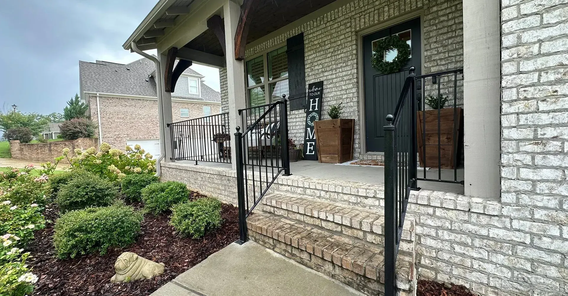 Brick house porch with black railing, potted plants, and front steps beside a garden path