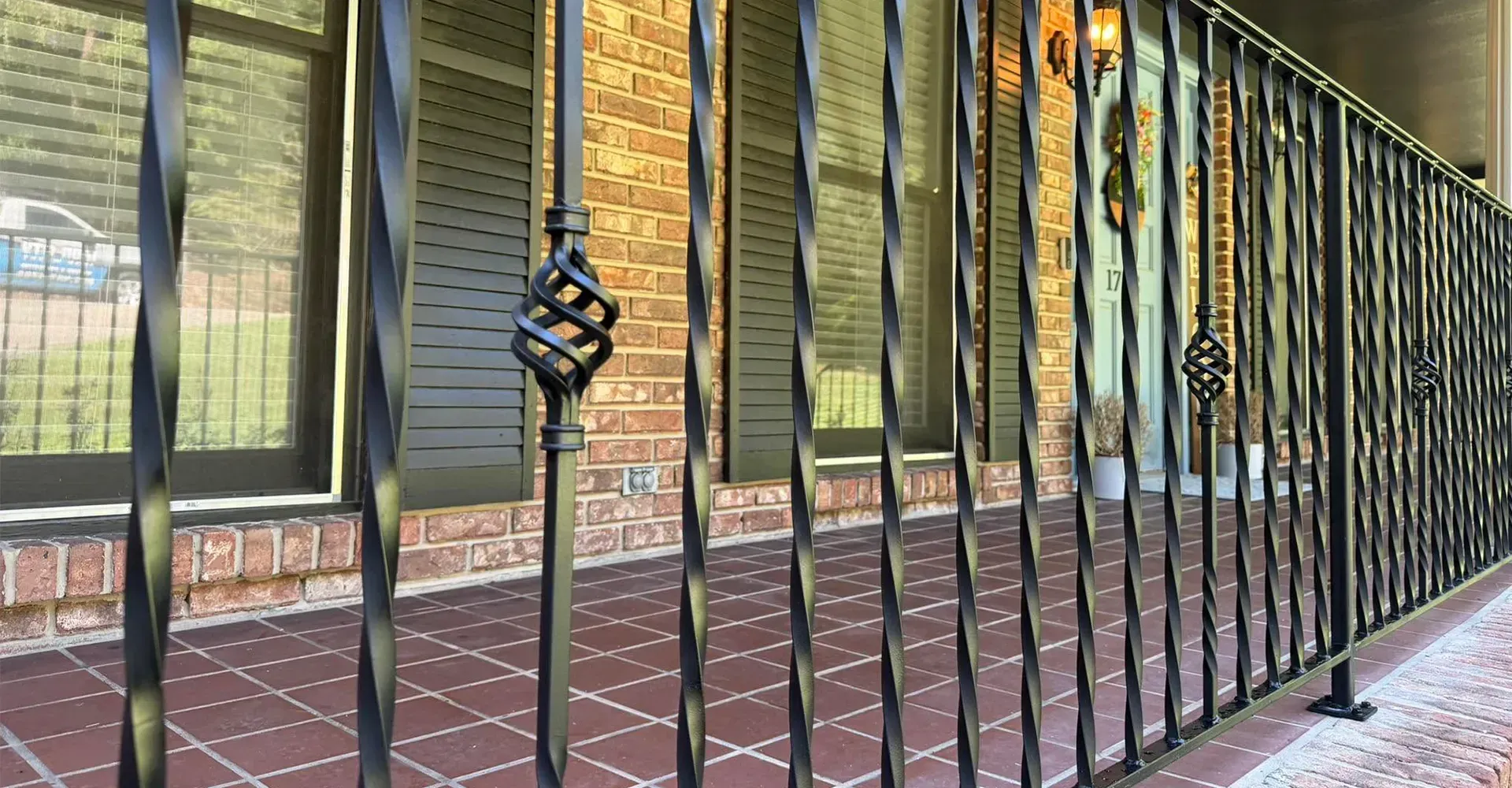 Black metal porch railing in front of a brick house with windows and green shutters