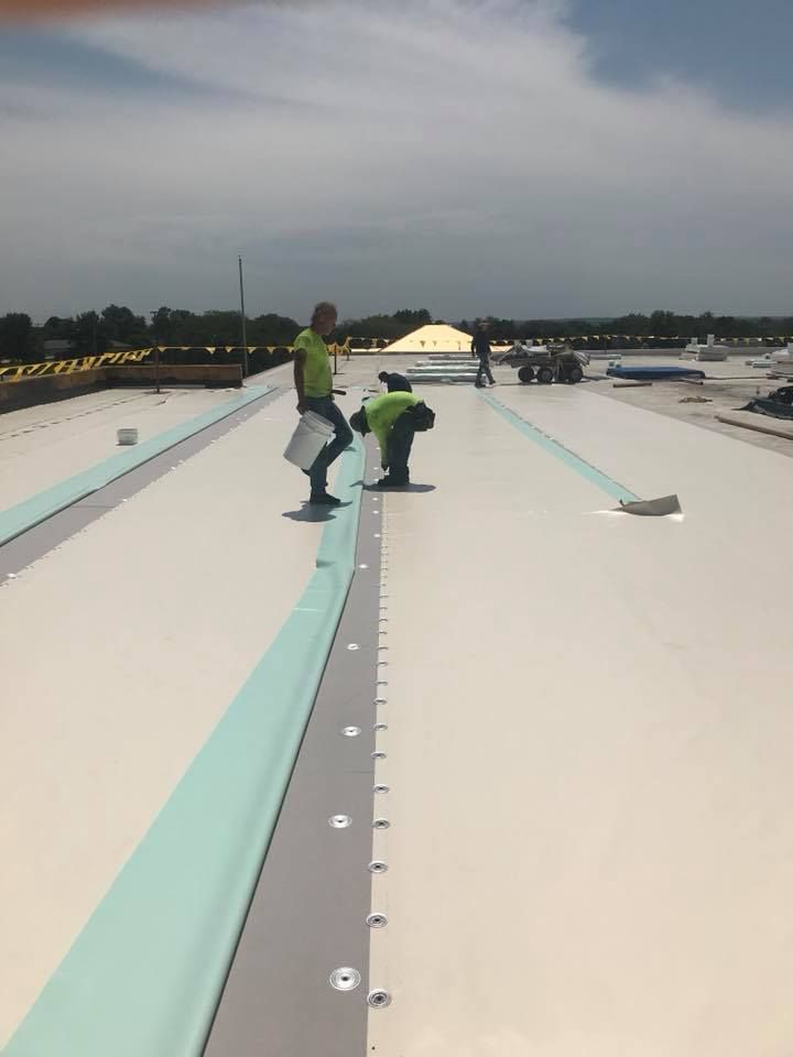 Two workers in green safety gear on a white rooftop near a long seam, with a city skyline in the distance