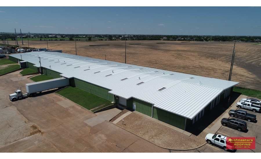 Aerial view of a large white metal warehouse beside a dirt lot with parked vehicles.